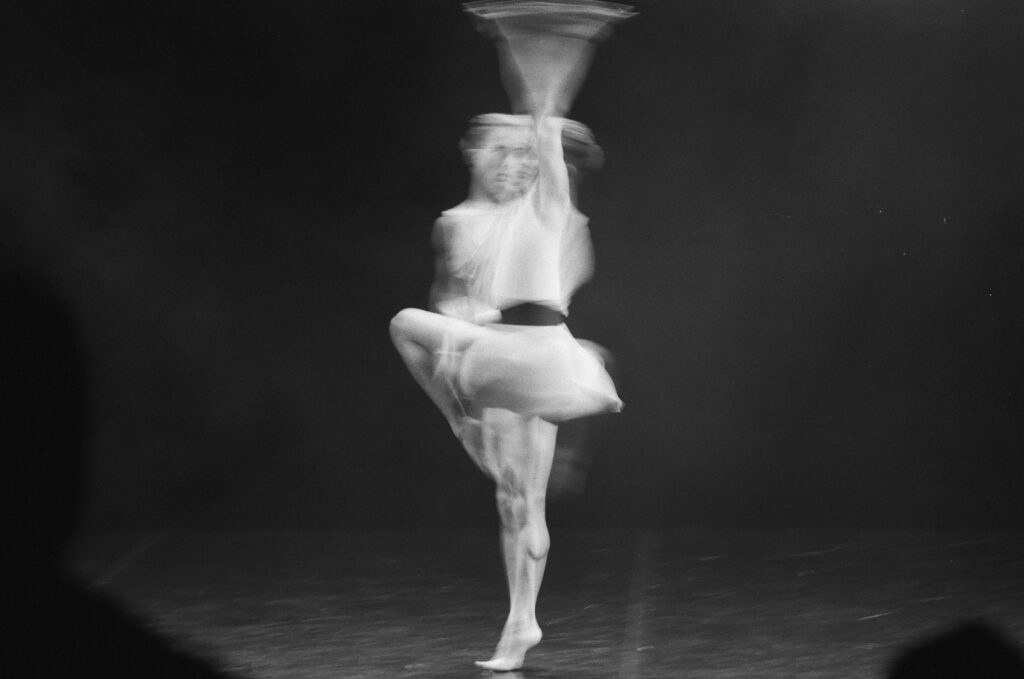 A black and white photograph capturing the motion of a ballet dancer in a graceful pose.